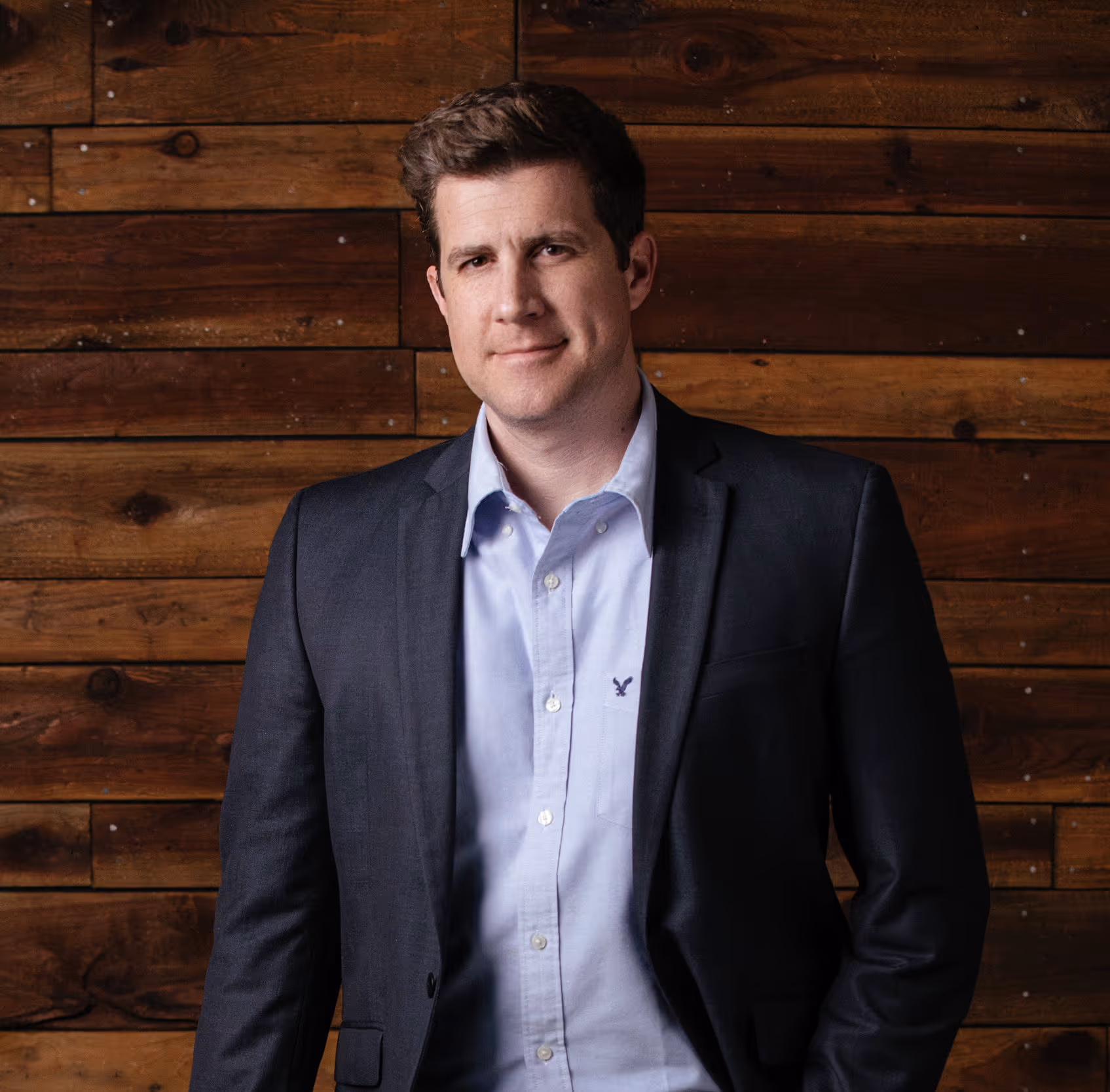 Man in a dark blazer and light blue shirt standing against a wooden plank wall.