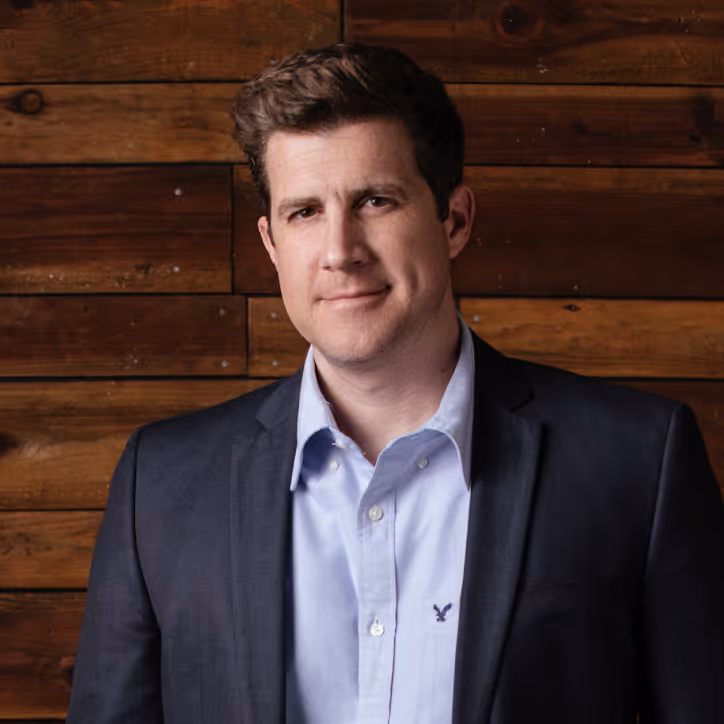 Man with short brown hair wearing a dark blazer and light blue button-up shirt standing in front of a wooden wall.